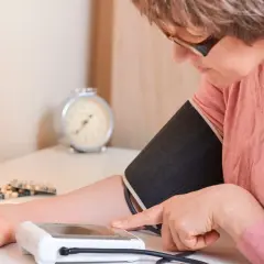 Kobieta mierzy ciśnienie, zmagając się z **wysokim ciśnieniem**. Na stole leżą leki.