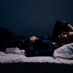Kobieta w piżamie leży na łóżku, z ręką na czole. Ciemne otoczenie sugeruje noc, a jej wyraz twarzy może wskazywać na trudności ze snem, być może związane z narkolepsją.
