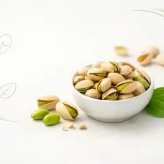 Misternie ułożone pistacje w miseczce, obok kilka luźnych orzechów i liści. Ikony serca, talerza i liścia sugerują, czy pistacje są zdrowe.