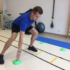 Mężczyzna w niebieskiej koszulce ćwiczy z drabinką koordynacyjną, zielonymi pachołkami i matą. W tle rower stacjonarny i drabinki.