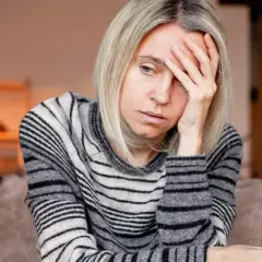Kobieta z blond włosami, w swetrze w paski, trzyma dłoń na czole, wyraźnie cierpiąc z powodu silnego bólu głowy.