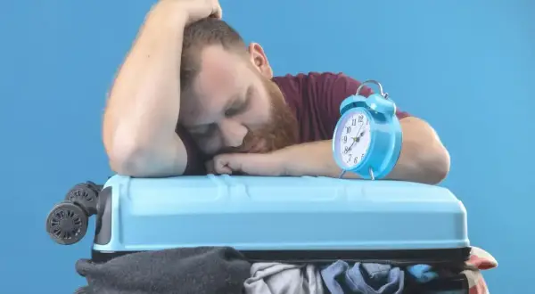 Zmęczony podróżnik śpi na walizce, budzik obok sygnalizuje jetlag.