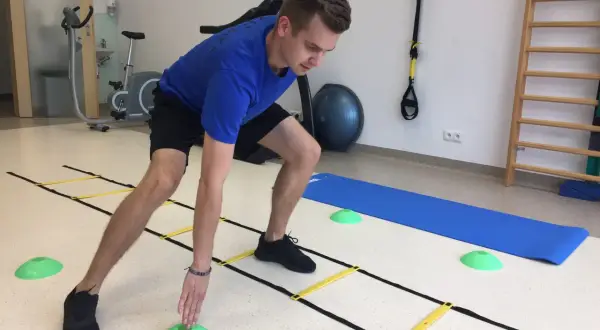 Mężczyzna w niebieskiej koszulce ćwiczy z drabinką koordynacyjną, zielonymi pachołkami i matą. W tle rower stacjonarny i drabinki.