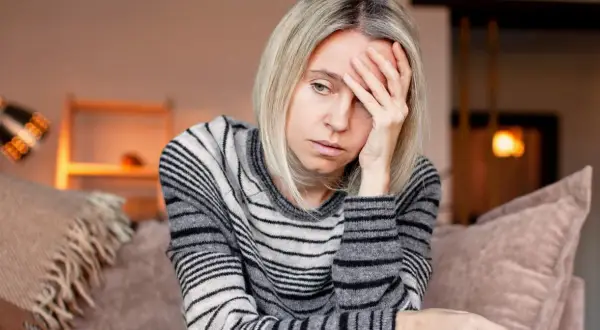 Kobieta z blond włosami, w swetrze w paski, trzyma dłoń na czole, wyraźnie cierpiąc z powodu silnego bólu głowy.