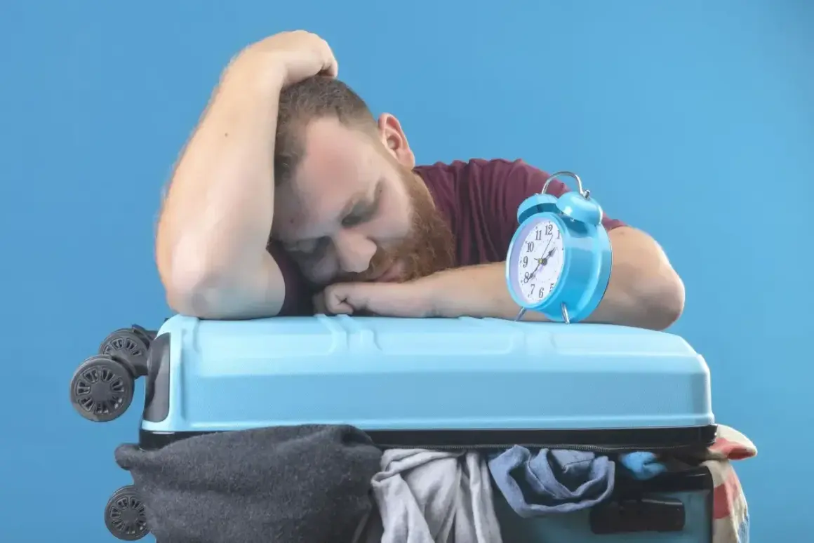 Zmęczony podróżnik śpi na walizce, budzik obok sygnalizuje jetlag.
