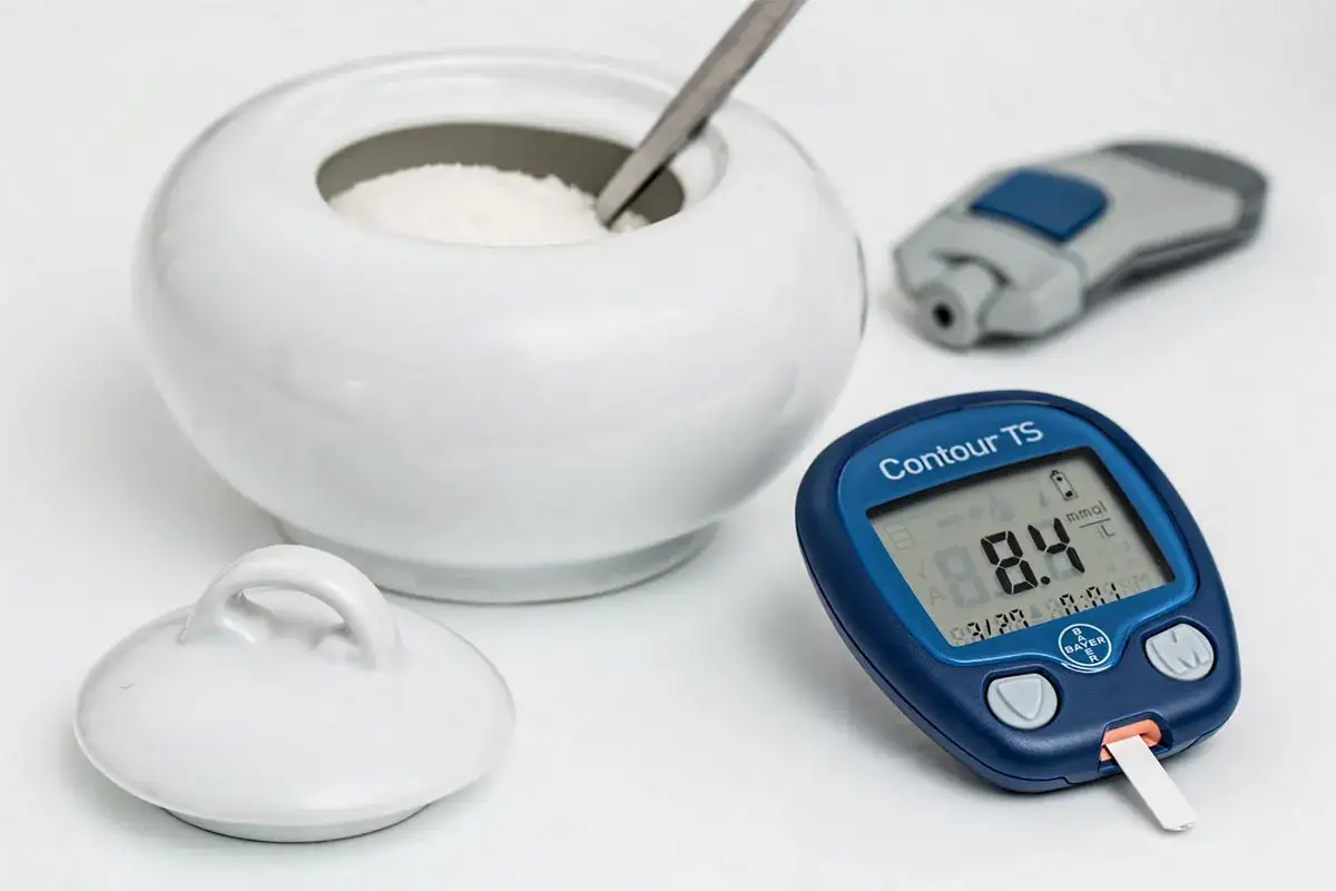Glukometr pokazuje 8.4 mmol/L. Obok cukiernica i nakłuwacz. Leczenie cukrzycy, np. glucophage, wymaga kontroli.