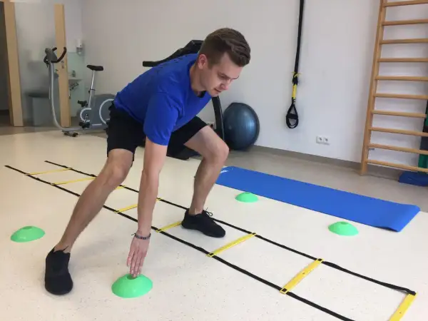 Mężczyzna w niebieskiej koszulce ćwiczy z drabinką koordynacyjną, zielonymi pachołkami i matą. W tle rower stacjonarny i drabinki.