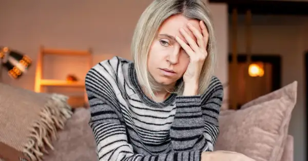 Kobieta z blond włosami, w swetrze w paski, trzyma dłoń na czole, wyraźnie cierpiąc z powodu silnego bólu głowy.